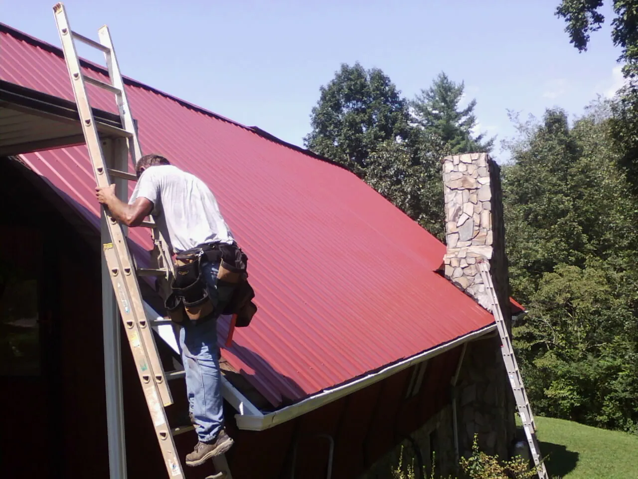 Roofer on a ladder beside a red metal roof and stone chimney — Soffit & Fascia Repair by Roofing Brothers in Defuniak Springs